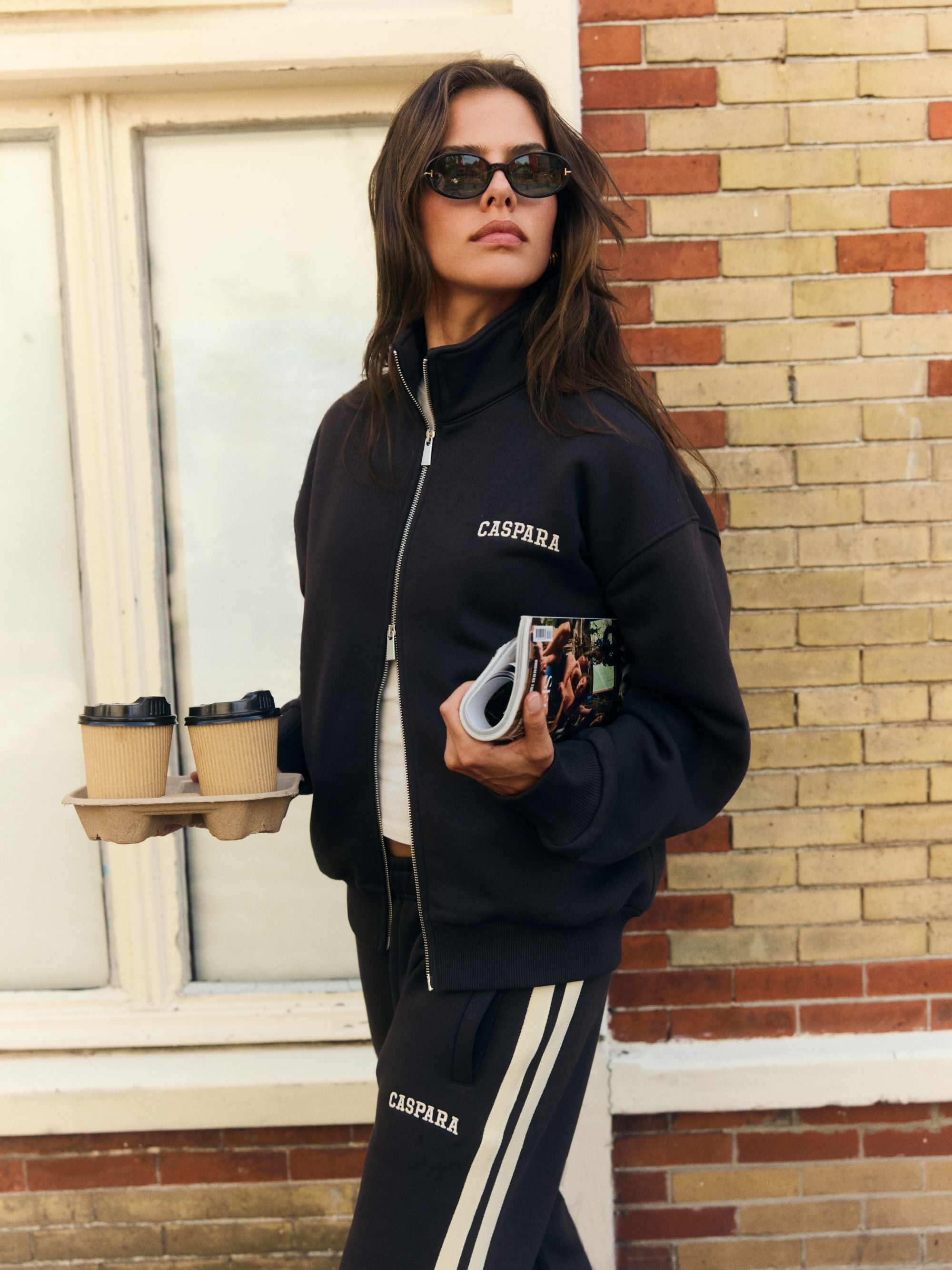 Woman wearing an espresso CASPARA funnel zip sweatshirt and sunglasses holding coffees outdoors.